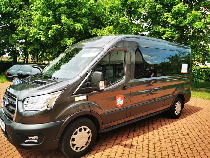 Pokaż zdjęcie: Srebrny bus stojący na parkingu z naklejkami z programu door-to -door. W tle drzewa