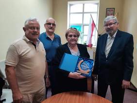 Pokaż zdjęcie: W gabinecie znajduje się jedna kobieta i trzech mężczyzn. Kobieta trzyma w rękach statuetkę, którą zamierza wręczyć. Wszyscy są uśmiechnięci i patrzą w obiektyw aparatu. 