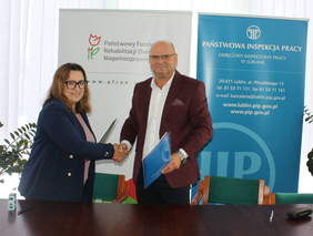 Pokaż zdjęcie: Mężczyzna i kobieta stojący na tle rollupów PFRON oraz Inspekcji Pracy gratulują sobie trzymając się za ręce.