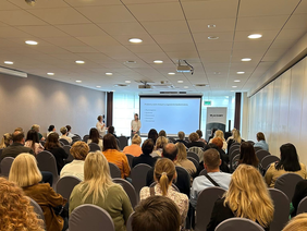 Pokaż zdjęcie: sala konferencyjna wypełniona ludźmi, prezentacja o zagrożeniu bezdomnością młodzieży, slajd z pięcioma punktami, dwóch prelegentów, ekran i baner z napisem „WŁĄCZAMY”.