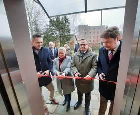 Pokaż zdjęcie: na zdjęciu budynek z zamontowaną windą po środku stoją osoby i przecinają  czerwoną wstęgę 