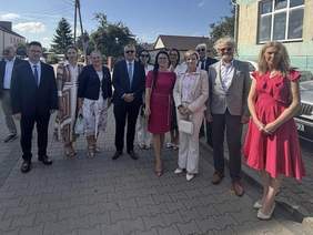 Pokaż zdjęcie: Grupa kobiet i mężczyzn stoi wspólnie na ulicy podczas słonecznego dnia. Uczestnicy są elegancko ubrani, pozują do zdjęcia przed uroczystością jubileuszową Domu Pomocy Społecznej w Brańsku. W tle widoczne są budynki mieszkalne .