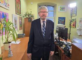 Pokaż zdjęcie: Dyrektor Łódzkiego Oddziału PFRON stoi w korytarzu przy sekretariacie. Po prawej stronie widać komputery i inne urządzenia biurowe. Na ścianach wiszą kolorowe obrazki. 