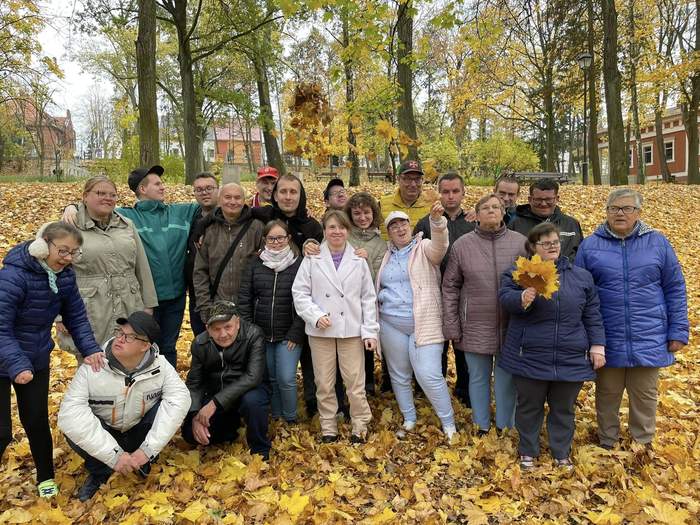 Pokaż zdjęcie: Grupa osób z niepełnosprawnościami stoi obok siebie na tle jesiennej aury, są uśmiechnięci i zadowoleni