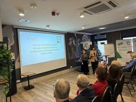 Pokaż zdjęcie: Przy ekranie stoją dwie panie, które wygłaszają prelekcję. 