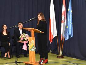 Na środku sali, przy mównicy przedstawicielka PFRON odczytuje list gratulacyjny od  Dyrektor Kujawsko-Pomorskiego Oddziału PFRON Pani Aleksandy Gierej. Za nia stoją trzy flagi.