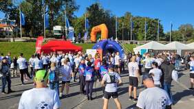 Pokaż zdjęcie: Tłum uczestników w koszulkach biegu „Po Prostu Bieg 2025” gromadzi się przy czerwonym i białych namiotach oraz niebieskiej bramce startowej. W tle widoczne flagi, drzewa i jasne niebo.