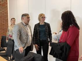 Pokaż zdjęcie: Przy stole stoi mężczyzna trzymający dłoń na krześle. Rozmawia z trzema kobietami stojącymi naprzeciw. Z tyłu stoi kobieta w okularach przysłuchująca się rozmowie.