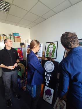 Pokaż zdjęcie: Trzy osoby stoją przed sztalugami z pracami i oglądają prace plastyczne.