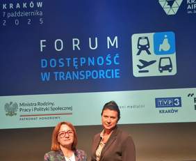 Pokaż zdjęcie: Dwie kobiety pozują na tle ekranu. Na ekranie na granatowym tle napis: Forum dostępności "Dostępność w transporcie" 