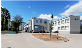 Pokaż zdjęcie: Zdjęcie przedstawia teren Ośrodka Wsparcia i Testów” w Białymstoku. W centralnej części widoczny jest nowoczesny budynek OWiT z wejściem po schodach oraz przeszkloną windą po lewej stronie. Na pierwszym planie znajduje się plac z drzewkami i hydrantem przeciwpożarowym, a po prawej stronie widoczny jest główny budynek szkoły z logo ZSZ5. W tle można dostrzec inne budynki szkolne oraz drzewa.