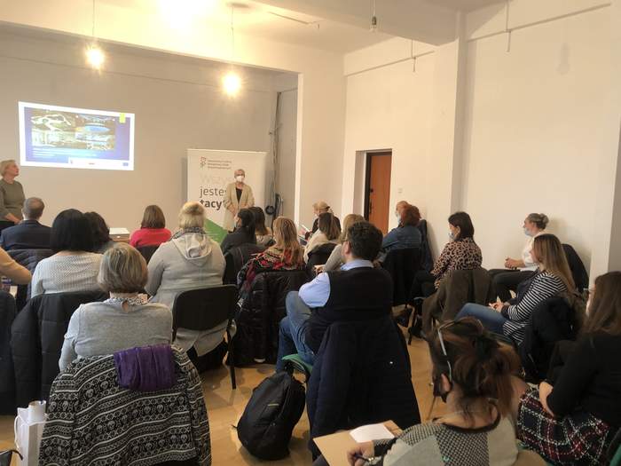 Pokaż zdjęcie: Fragment sali, od tyłu siedzący uczestnicy patrzą na wyświetlaną na środku ściany prezentację . Z przodu Sali przodem do słuchaczy stoją Eksperci PFRON: w lewym rogu Joanna Hryniewicka. W prawej części sali Teresa Saczuk-Grabowska