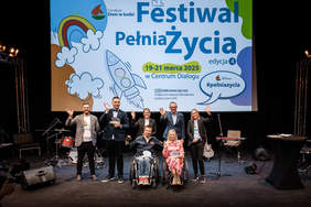 Pokaż zdjęcie: Na scenie znajduje sie siedem osób: trzy kobiety i czterech mężczyzn. Dwie osoby siedzą na ręcznych wózkach inwalidzkich, a pozostali stoją. Wszyscy się uśmiechają i machają dłońmi. 