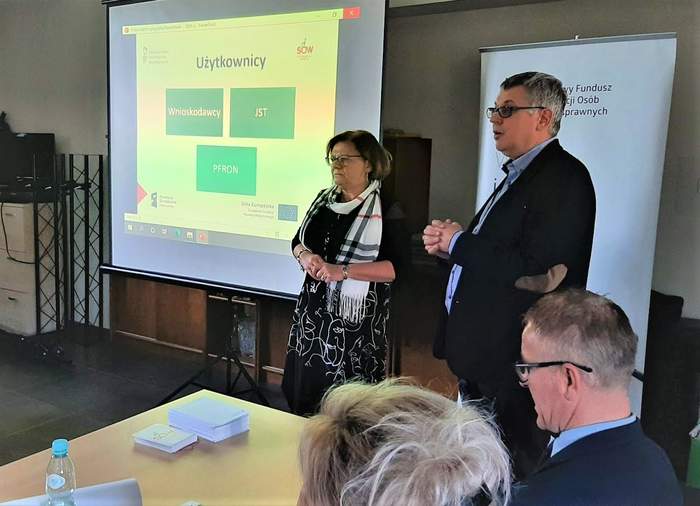 Pokaż zdjęcie: Na środku sali stoi Dyrektor i pracownik Oddziału, przedstawiając zalety systemu SOW, w tkle widoczny ekran, z wyświetlonym panelem, dotyczącym przedmiotowego systemu  