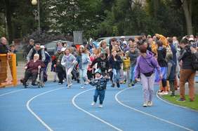 Pokaż zdjęcie: stadion w Wieruszowie po środku biegnie dużo osób  po lewej osoba na wózku, z przodu biegnie dziecko w wieku około 6 lat z tyłu widać drzewa , za barierkami stoją ludzie i kibicują uczestnikom biegu.