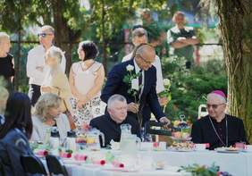 Pokaż zdjęcie: W plenerze przy stołach siedzą zaproszeni goście, na pierwszym planie kobieta i dwóch księży, stoły stoją w otoczeniu zieleni  