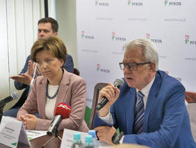 Pokaż zdjęcie: Zbliżenie postaci. Dwie osoby siedzą przy stole. Jedna z nich mówi do mikrofonu. W tle tłumacz języka migowego.