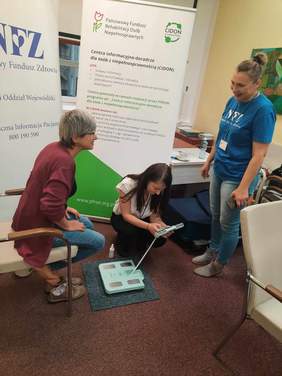 Pokaż zdjęcie: W pokoju znajdują się trzy kobiety. Jedna z pań siedzi na krześle i ma zdjęte buty. Druga pani kuca i wykonuje badanie analizy składu ciała siedzącej kobiety. Obok nich stoi trzecia pani i uśmiecha się szeroko. 