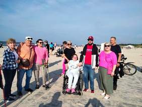 Pokaż zdjęcie: Grupa osób znajduje się na plaży. Niektórzy siedzą na wózkach inwalidzkich, inni trzymają kijki bądź laski. W tle widać nadmorski horyzont. 