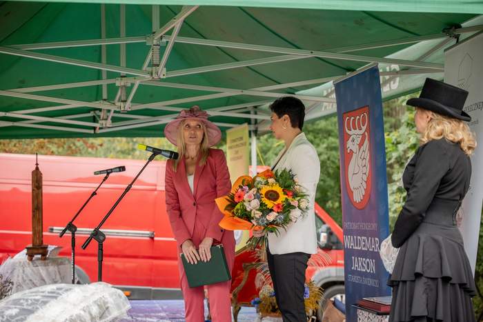 Pokaż zdjęcie: Na scenie stoją trzy panie, kobieta w malinowym garniturze i kapeluszu składa życzenia, druga z kobiet trzyma bukiet kwiatów. Tyłem stoi kobieta w pięknej czarnej sukni.