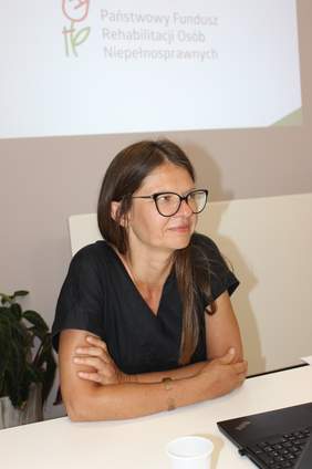 Kobieta siedzi przy stole podczas spotkania, na ekranie w tle widoczne jest logo PFRON.