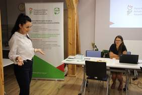 Kobieta wita zebranych na spotkaniu informacyjnym PFRON, a prelegentka siedzi przy stole.