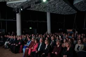 Pokaż zdjęcie: Widownia konferencyjna wypełniona uczestnikami siedzącymi w rzędach i zwróconymi w stronę sceny. Sala z nowoczesnym oświetleniem, dekoracyjnym sufitem oraz kolumnami. Na ścianie widoczny wyświetlacz z odliczaniem czasu.