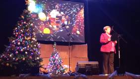 Pokaż zdjęcie: Na scenie przy mikrofonie stoi kobieta ubrana w różową marynarkę i czarne spodnie. Na scenie stoją dwie udekorowane świątecznie choinki.
