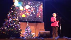 Pokaż zdjęcie: Na scenie przy mikrofonie stoi kobieta ubrana w różową marynarkę i czarne spodnie. Na scenie stoją dwie udekorowane świątecznie choinki.