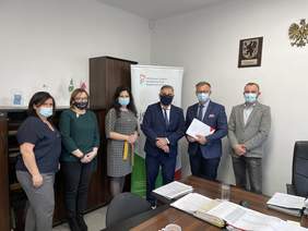 Pokaż zdjęcie: W biurze z ciemnymi meblami, jasnymi ścianami stoi trzech mężczyzn i trzy kobiety, za nimi stoi baner z logo PFRON. Jeden z mężczyzn ubrany w granatowy garnitur biała koszulę i krawat trzyma biała teczkę z logo PFRON.