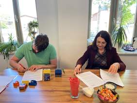 Pokaż zdjęcie: Przy stole siedzi kobieta ubrana w czarną marynarkę  i mężczyzna ubrany w koszulkę polo, podpisują dokumenty. Na biurku leżą pieczątki. Za nimi na parapetach stoją kwiaty w doniczkach.