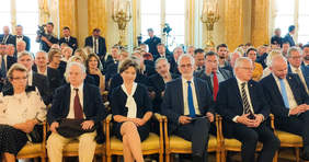 Pokaż zdjęcie: Kilkadziesiat osób siedzi na krzesłach w kolorze złotym w sali zamku. 