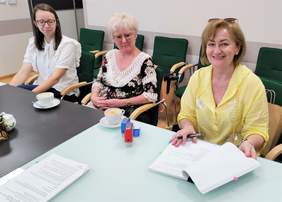 Pokaż zdjęcie: Przy stole siedzą trzy kobiety. Jedna z nich podpisuje dokumenty.