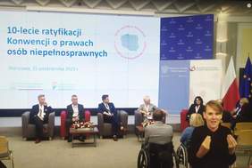 Pokaż zdjęcie: Zdjęcie konferencji z tyłu duży ekran z napisem: Konferencja z okazji 10-lecia ratyfikacji Konwencji o prawach osób niepełnosprawnych, przed ekranem w fotelach siedzi czterech prelegentów, z boku na fotelu kobieta z mikrofonem w niebieskim garniturze. W prawym dolnym rogu zdjęcia widać kobiecą postać tłumacza języka migowego