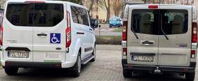 Pokaż zdjęcie: Dwa busy stoją zaparkowane na chodniku. Na masce jednego z nich umieszczona jest naklejka informująca, że pojazd dostosowany został do przewozu osób poruszających się na wózku. W tle dostrzec można ulicę, bloki oraz samochody osobowe.