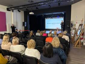 Pokaż zdjęcie: Grupa kilkunastu osób siedzi w sali konferencyjnej z wyniesioną sceną, na której znajduje się mężczyzna trzymający w dłoni mikrofon. Po lewej stronie sali widoczny jest drugi mężczyzna, który robi zdjęcia aparatem fotograficznym. Po prawej stronie możemy dostrzec baner powiatu gnieźnieńskiego oraz trzy sztalugi, na których zostały umieszczone tablice informacyjne.