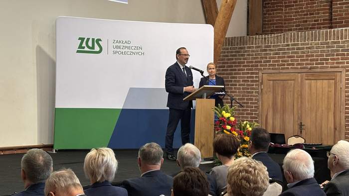 Pokaż zdjęcie: Mężczyzna na tle baneru ZUS przemawia do mikrofonu. Obok niego stoi kobieta. W sali znajduje się także grupa kilkunastu osób, które siedzą i przysłuchują się wystąpieniu.