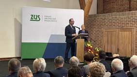 Pokaż zdjęcie: Mężczyzna na tle baneru ZUS przemawia do mikrofonu. Obok niego stoi kobieta. W sali znajduje się także grupa kilkunastu osób, które siedzą i przysłuchują się wystąpieniu.