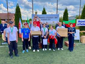 Pokaż zdjęcie: Grupa kilkunastu osób stoi na boisku. Wśród nich są przedstawiciele instytucji i zawodnicy z otrzymanymi medalami.