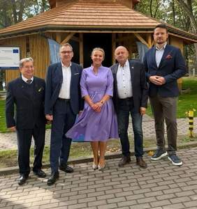 Pięcioosobowa grupa osób pozuje przed drewnianą, stylizowaną tężnią solankową w otoczeniu zieleni. Na pierwszym planie stoją trzy osoby w garniturach, kobieta w fioletowej sukience oraz mężczyzna w marynarce. W tle widoczny jest budynek tężni z charakterystycznym drewnianym dachem.