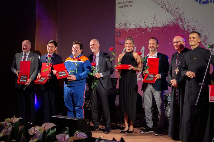 Pokaż zdjęcie: Scena na której stoi 8 osób w rzędzie, trzymają w ręku uroczyste podziękowania oraz pojedyncze kwiaty