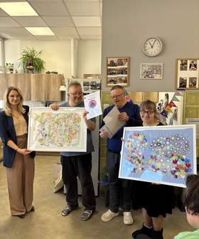 Grupa osób prezentuje kolorowe prace plastyczne i dyplomy w pomieszczeniu biurowym. Na pierwszym planie widoczne duże obrazy: abstrakcja oraz kwiecista kompozycja na niebieskim tle.