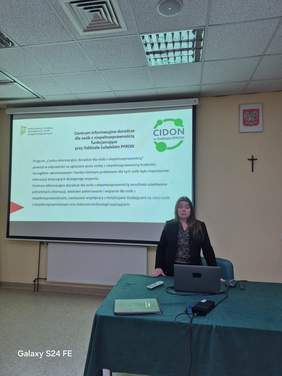 Pokaż zdjęcie: Kobieta prelegent stoi przy biurku, w tle slajd z prezentacji.