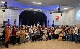 Pokaż zdjęcie: Pamiątkowe zdjęcie grupowe uczestników wydarzenia w jasnej auli z drewnianą podłogą i sceną w tle. Liczna grupa dorosłych i młodzieży (w tym osoby na wózkach) pozuje w szerokim półokręgu z uniesionymi rękami. Są ubrani w różnorodne, kolorowe stroje w stylu westernowo-farmerskim. Na ścianie po prawej stronie wisi herb z napisem „Zbąszyń”.