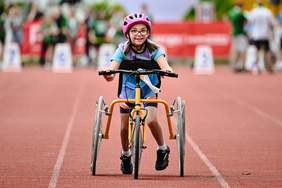 Pokaż zdjęcie: Frame Running  Przy specjalnie przystosowanym wózku trzykołowym biegnie uśmiechnięta dziewczynka ubrana na sportowo, w kasku.