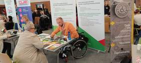 Punkt informacyjny Kujawsko-Pomorskiego Oddziału PFRON - przy stoliku siedzi meżczyżna - pracownik PFRON, a przed siedzi kobieta i rozmawiają. Mężczyzna pokazuje coś kobiecie w laptopie. Na stoliku leżą ulotki, broszury i informatory oraz materiały reklamowe. w tle widać banery PFRON CIDON