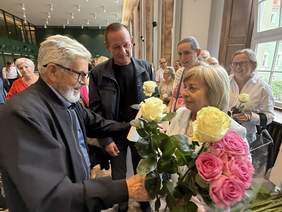 Pokaż zdjęcie: Grupa osób podczas uroczystości. W centralnym punkcie starszy mężczyzna wręcza Barbarze Jaskierskiej bukiet kwiatów. Pani Jaskierska trzyma w rękach bukiet różowych i kremowych róż. W tle uśmiechnięci uczestnicy wydarzenia.