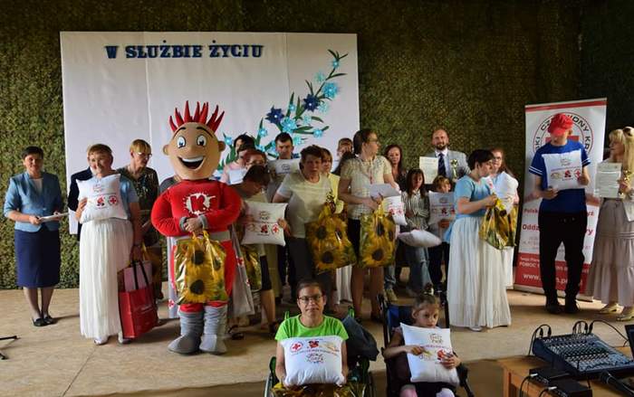 Pokaż zdjęcie: Na scenie stoi duża grupa osób, osoby starsze i młodsze, trzymające w rękach dyplomy i nagrody-poduszki. Pomiędzy laureatami stoi duża pluszowa maskotka imitująca kroplę krwi. 