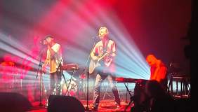 Pokaż zdjęcie: Dwóch mężczyzn stojących przy stojaku z mikrofonem, jeden trzyma gitarę w ręku. Obok nich siedzi mężczyzna przy syntezatorze. W tle widać perkusję oraz gitarę. Wszystko to odbywa się na scenie