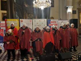 Pokaż zdjęcie: W kościele stoją artyści, którzy śpiewają kolędy i pastorałki. Wszyscy są ubrani na czerwono. 